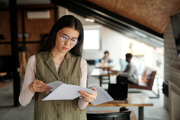 Wall Mural - Young serious businesswoman in eyeglasses looking through contract or other financial documents while standing in front of camera in office