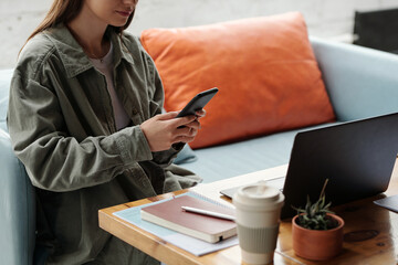 Wall Mural - Cropped shot of young businesswoman in grey shirt texting in smartphone while sitting on comfortable couch by workplace in front of laptop