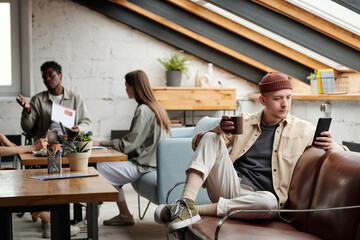 Aufkleber - Young serious businessman with cup of coffee and mobile phone scrolling through online photos or posts while sitting on leather sofa in office
