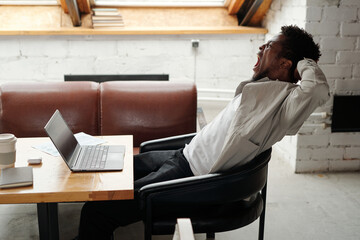 Aufkleber - Side view of young yawning businessman with hands behind his head sitting in armchair by workplace with laptop and having minute of rest