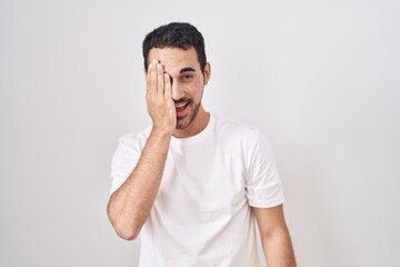 handsome hispanic man standing over white background covering one eye with hand, confident smile on 