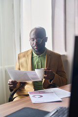 Leinwandbilder - Young serious businessman with cup of coffee looking through financial documents while sitting by workplace in front of camera in office