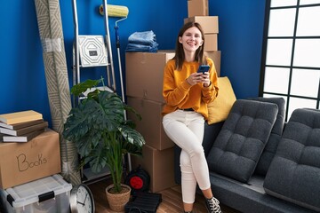 Poster - Young blonde woman using smartphone sitting on sofa at new home