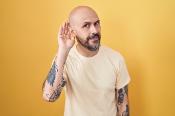 Poster - Hispanic man with tattoos standing over yellow background smiling with hand over ear listening an hearing to rumor or gossip. deafness concept.