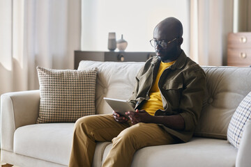 Fototapete - Young African American male freelancer with tablet sitting on soft comfortable couch in living room and looking through online data