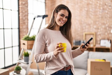 Wall Mural - Young beautiful hispanic woman using smartphone drinking coffee at new home