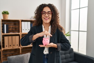 Sticker - Young hispanic woman holding piggy bank winking looking at the camera with sexy expression, cheerful and happy face.