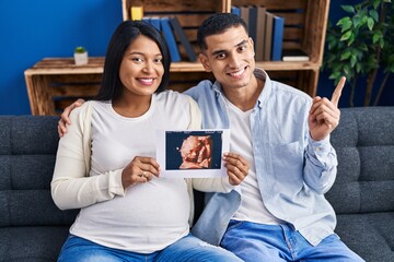 Sticker - Young hispanic couple expecting a baby sitting on the sofa showing baby ultrasound smiling happy pointing with hand and finger to the side