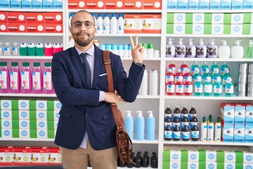 Poster - Hispanic man with beard working as salesman at pharmacy drugstore smiling with happy face winking at the camera doing victory sign. number two.