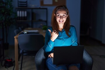 Sticker - Brunette woman working at the office at night smiling with happy face looking and pointing to the side with thumb up.