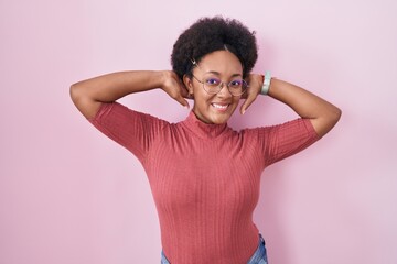 Poster - Beautiful african woman with curly hair standing over pink background relaxing and stretching, arms and hands behind head and neck smiling happy