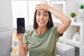 Canvas Print - Young brunette woman holding smartphone showing blank screen stressed and frustrated with hand on head, surprised and angry face