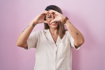 Wall Mural - Blonde woman standing over pink background smiling in love doing heart symbol shape with hands. romantic concept.