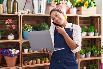 Wall Mural - Young beautiful hispanic woman florist talking on smartphone using laptop at florist