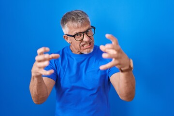 Sticker - Hispanic man with grey hair standing over blue background shouting frustrated with rage, hands trying to strangle, yelling mad