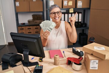 Wall Mural - Middle age woman working at small business ecommerce holding dollars smiling happy pointing with hand and finger to the side