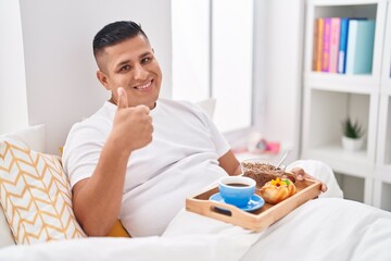 Poster - Young hispanic man eating breakfast in the bed smiling happy and positive, thumb up doing excellent and approval sign