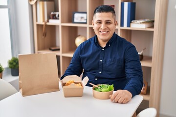 Canvas Print - Hispanic young man eating take away food looking positive and happy standing and smiling with a confident smile showing teeth