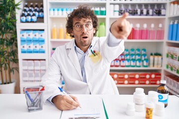 Wall Mural - Hispanic young man working at pharmacy drugstore pointing with finger surprised ahead, open mouth amazed expression, something on the front