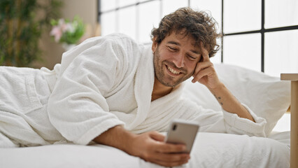 Wall Mural - Young hispanic man wearing bathrobe using smartphone smiling at bedroom
