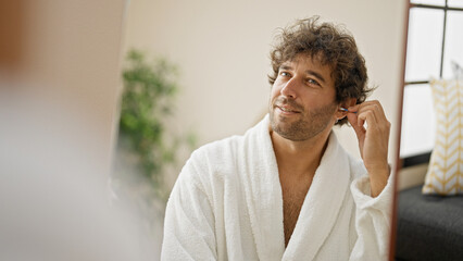 Young hispanic man wearing bathrobe cleaning ear with cotton swab looking on mirror at home