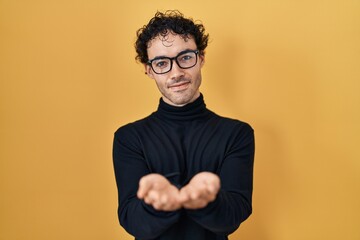 Wall Mural - Hispanic man standing over yellow background smiling with hands palms together receiving or giving gesture. hold and protection