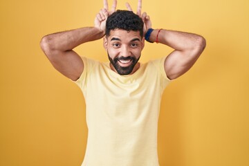 Canvas Print - Hispanic man with beard standing over yellow background posing funny and crazy with fingers on head as bunny ears, smiling cheerful