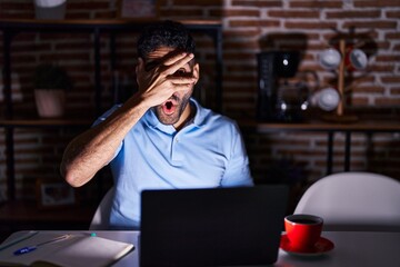 Wall Mural - Hispanic man with beard using laptop at night peeking in shock covering face and eyes with hand, looking through fingers with embarrassed expression.