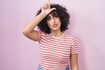 Poster - Young middle east woman standing over pink background making fun of people with fingers on forehead doing loser gesture mocking and insulting.