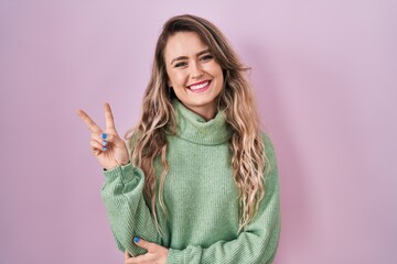 Sticker - Young caucasian woman standing over pink background smiling with happy face winking at the camera doing victory sign. number two.