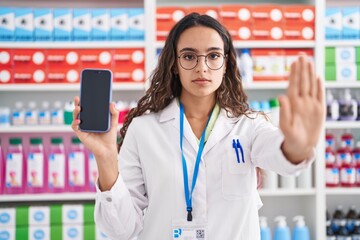 Wall Mural - Young hispanic woman working at pharmacy drugstore showing smartphone screen puffing cheeks with funny face. mouth inflated with air, catching air.