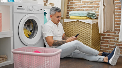 Wall Mural - Young hispanic man using smartphone waiting for washing machine at laundry room