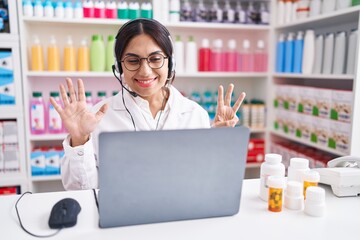 Wall Mural - Young arab woman working at pharmacy drugstore using laptop showing and pointing up with fingers number eight while smiling confident and happy.