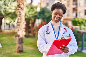 Wall Mural - African american woman wearing doctor uniform writing on clipboard at park