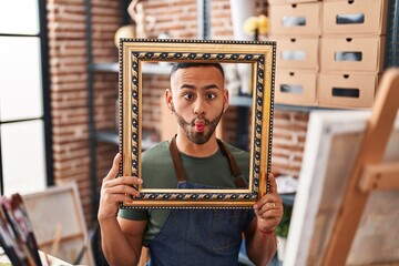 Poster - Young hispanic man sitting at art studio with empty frame making fish face with mouth and squinting eyes, crazy and comical.