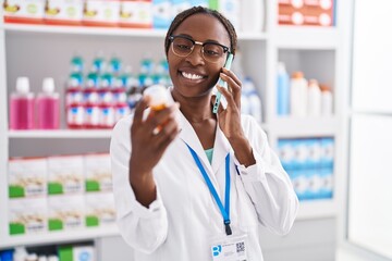 Canvas Print - African american woman pharmacist holding pills bottle talking on smartphone at pharmacy