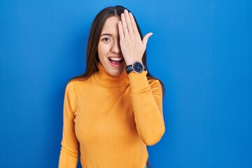 Poster - Young brunette woman standing over blue background covering one eye with hand, confident smile on face and surprise emotion.