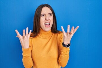 Sticker - Young brunette woman standing over blue background crazy and mad shouting and yelling with aggressive expression and arms raised. frustration concept.