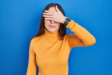 Poster - Young brunette woman standing over blue background covering eyes with hand, looking serious and sad. sightless, hiding and rejection concept