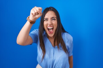 Poster - Young brunette woman standing over blue background angry and mad raising fist frustrated and furious while shouting with anger. rage and aggressive concept.