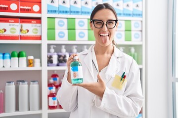 Sticker - Brunette woman working at pharmacy drugstore holding cough syrup sticking tongue out happy with funny expression.