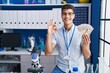 © Krakenimages.com - Young hispanic man working at scientist laboratory holding dollars doing ok sign with fingers, smiling friendly gesturing excellent symbol