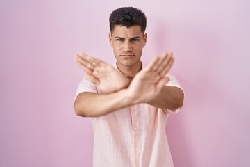 Sticker - Young hispanic man standing over pink background rejection expression crossing arms and palms doing negative sign, angry face