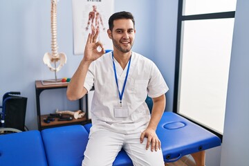 Poster - Young hispanic man with beard working at pain recovery clinic smiling positive doing ok sign with hand and fingers. successful expression.