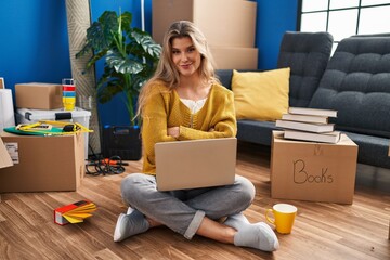 Canvas Print - Young woman sitting on the floor at new home using laptop happy face smiling with crossed arms looking at the camera. positive person.