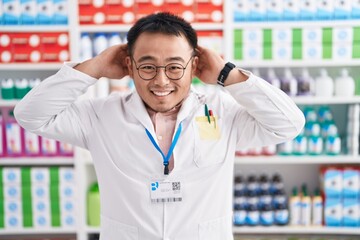 Poster - Chinese young man working at pharmacy drugstore relaxing and stretching, arms and hands behind head and neck smiling happy