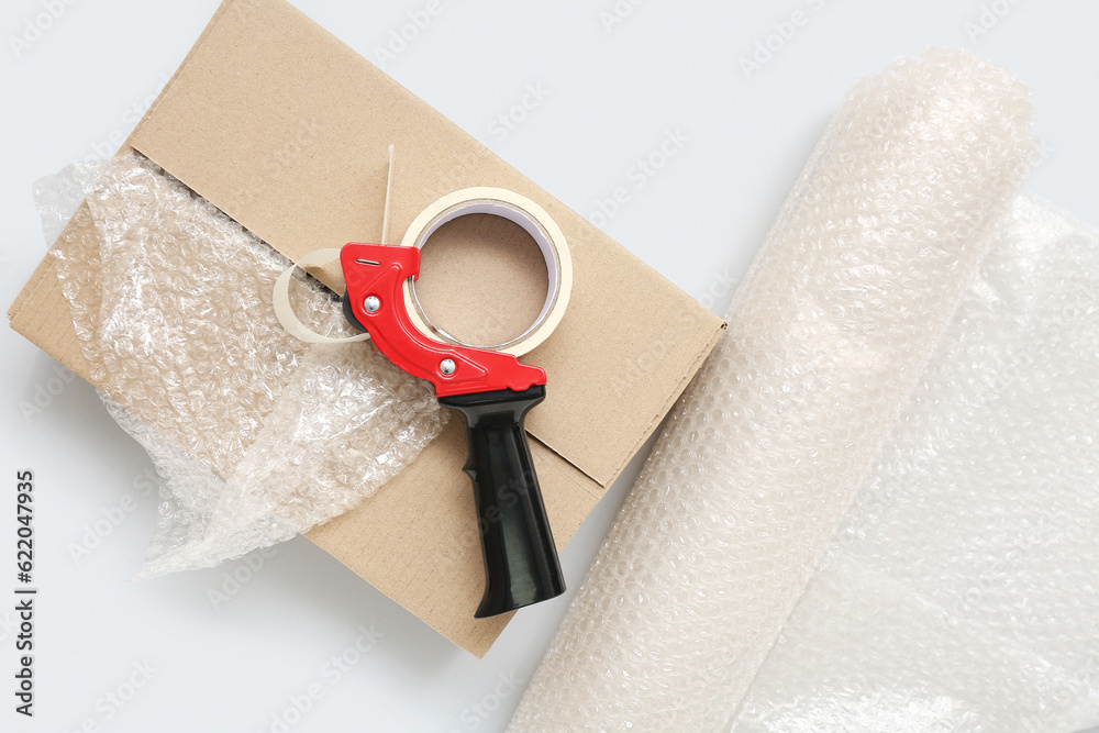 Packing tape dispenser, cardboard box and roll of bubble wrap on grey background