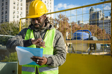 Wall Mural - young black engineer man with helmet and vest outdoors watching the time to go to eat and rest.