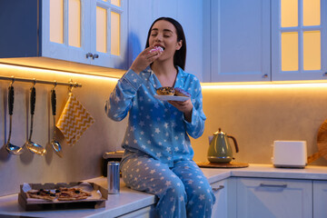 Wall Mural - Young woman eating donut in kitchen at night. Bad habit