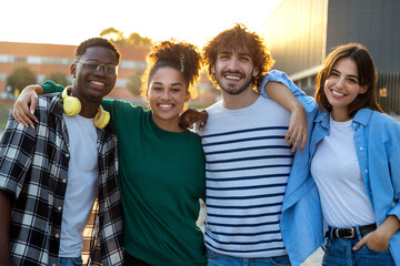 Group of happy and smiling multiracial friends embracing looking at camera standing outdoors during sunset.